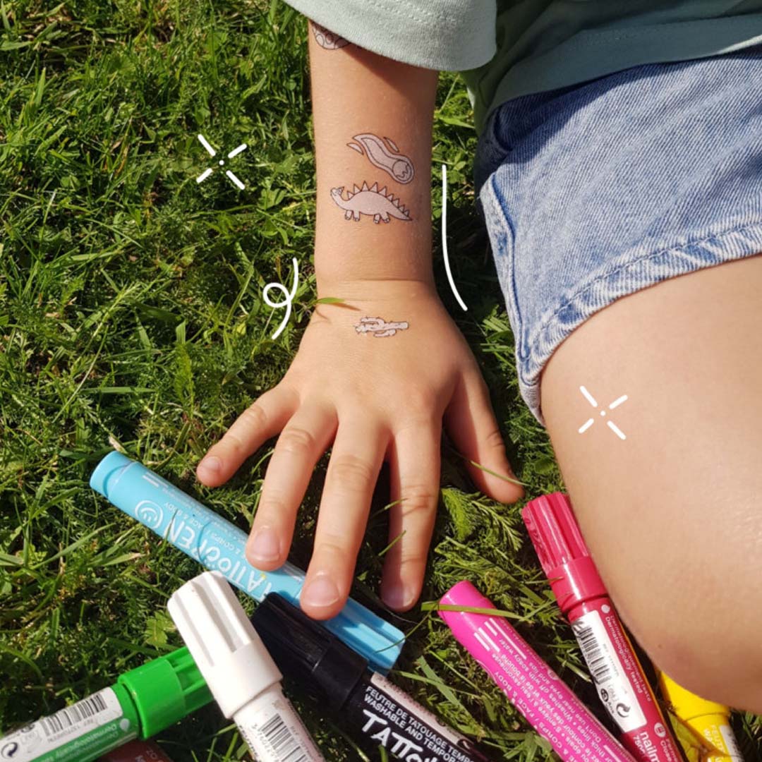 Een kinderarm met nailmatic dinosaurus tijdelijke tattoos op de pols en hand, omringd door kleurpotloden op gras.