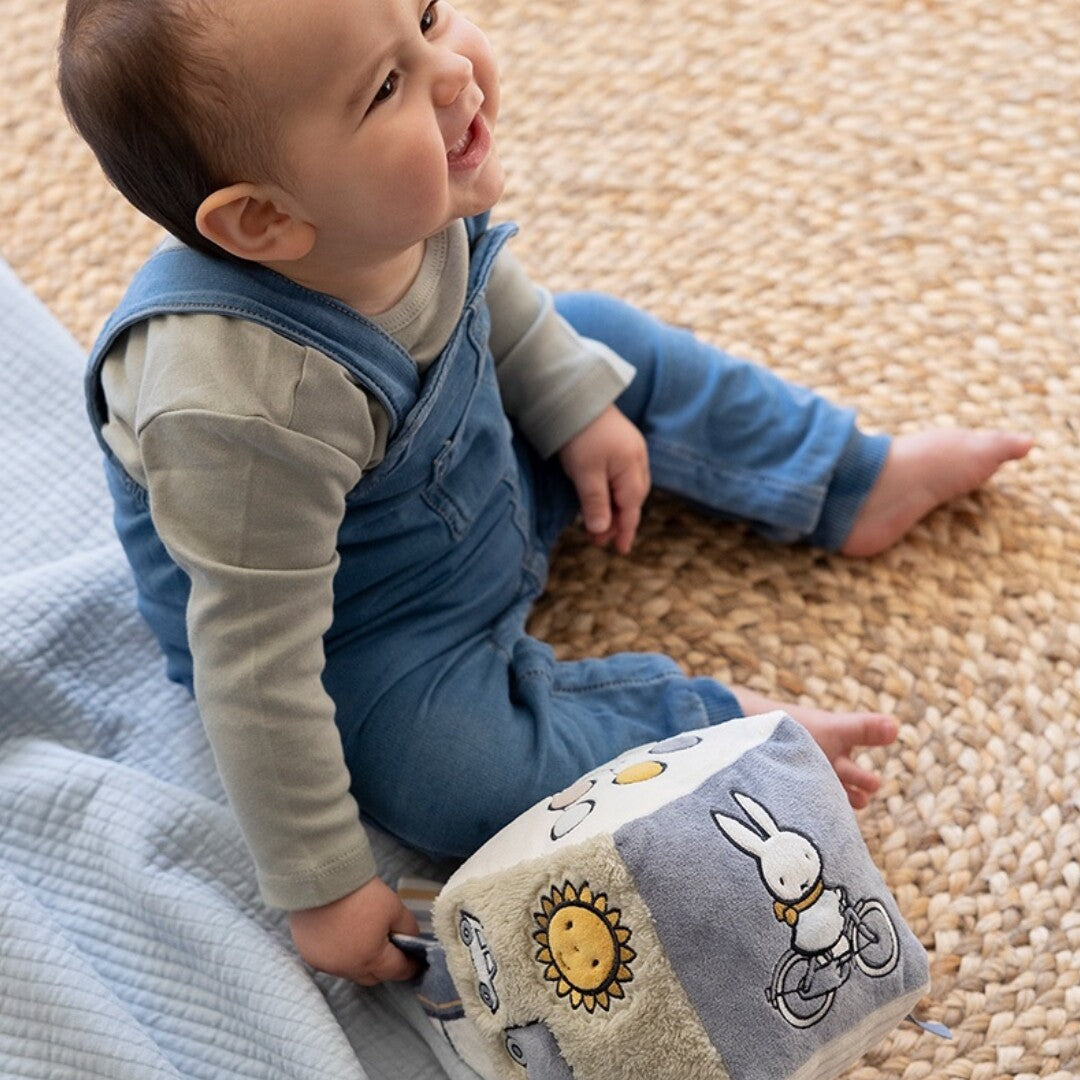 Een lachende baby zit op een mat naast de nijntje-miffy activiteitenkubus met Miffy op een fiets.