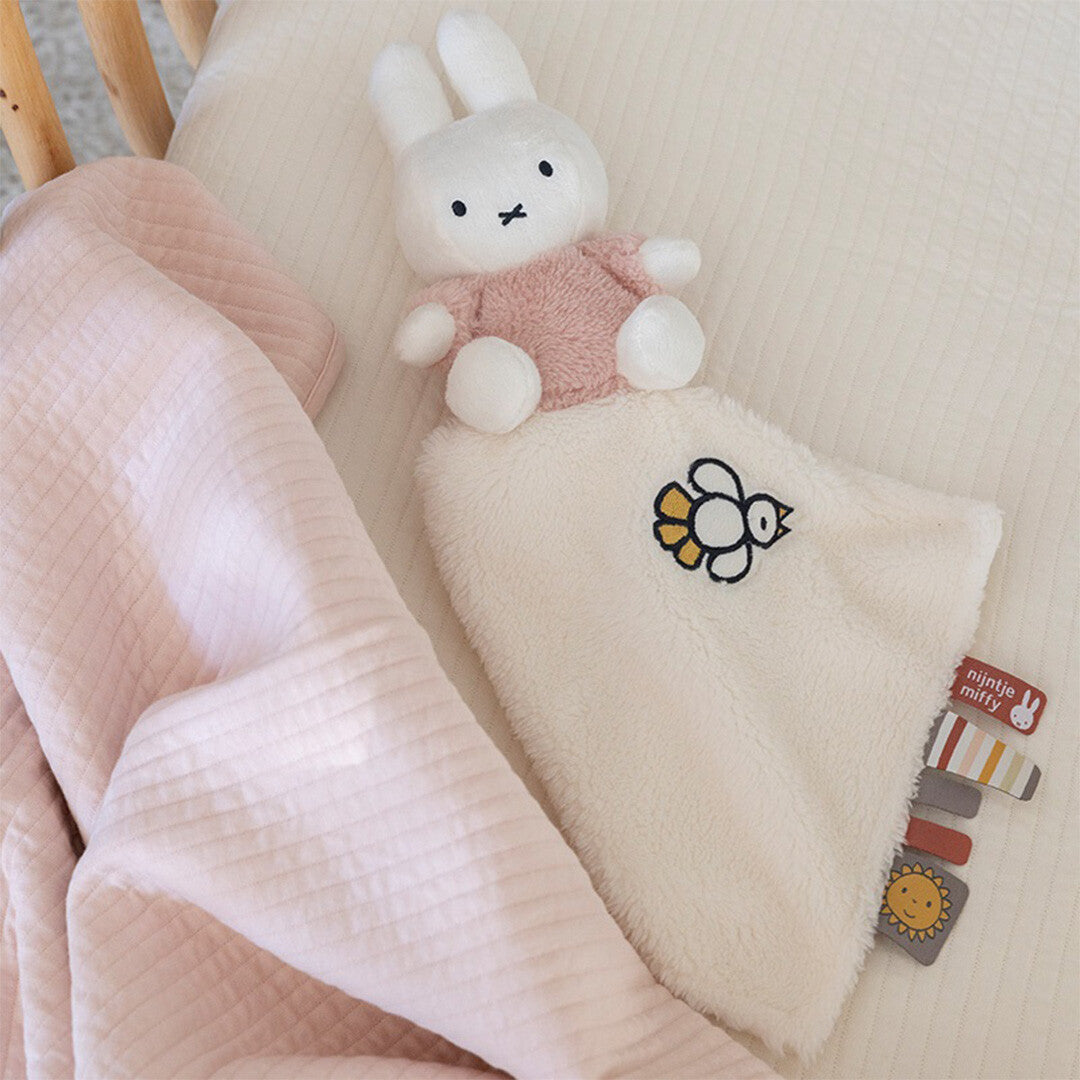 Het Nijntje-miffy pluchen knuffeldoekje met roze Nijntje konijn ligt in een wiegje naast een roze deken.