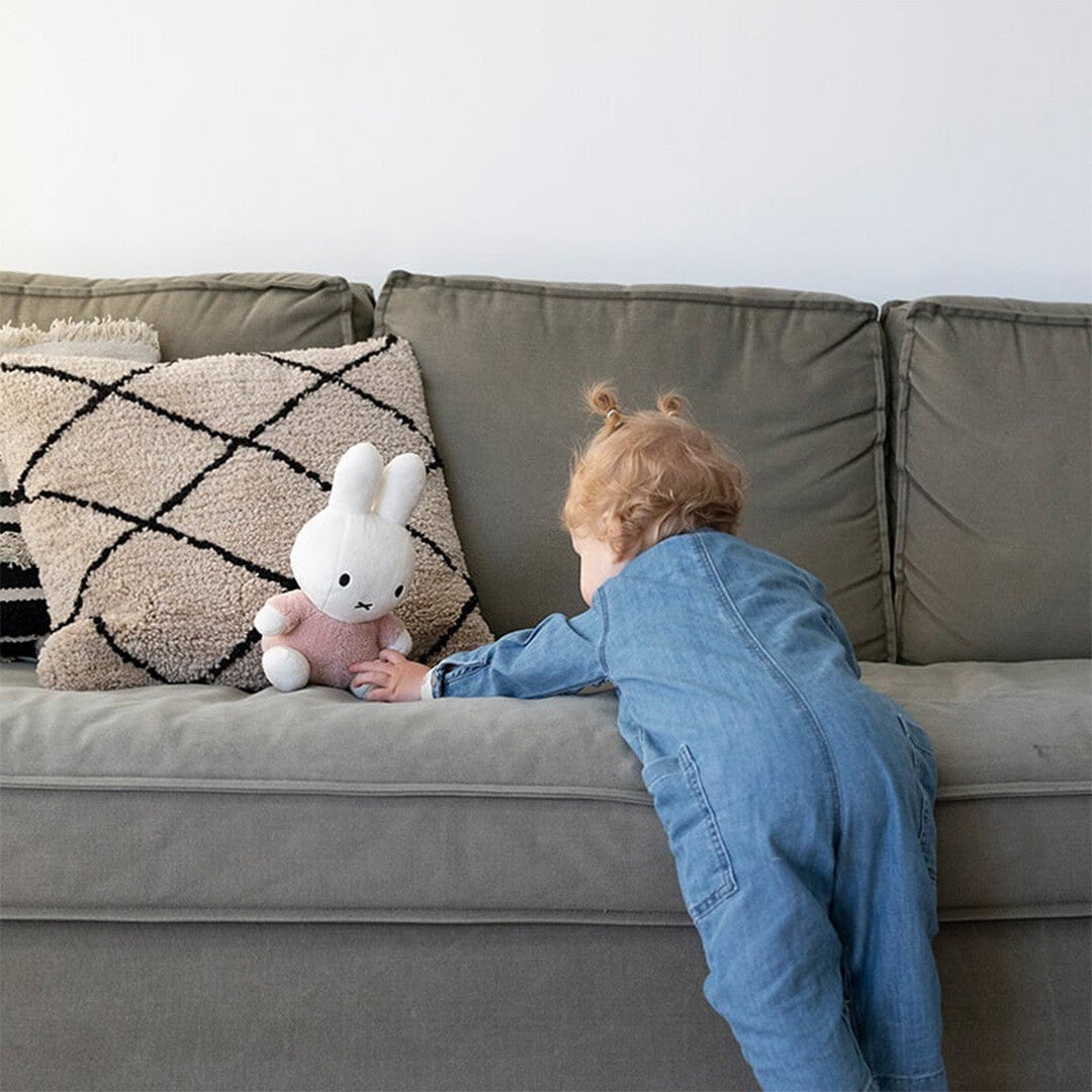Een kind in een spijkeroverall reikt naar het Nijntje-miffy knuffelkonijn op een grijze bank.
