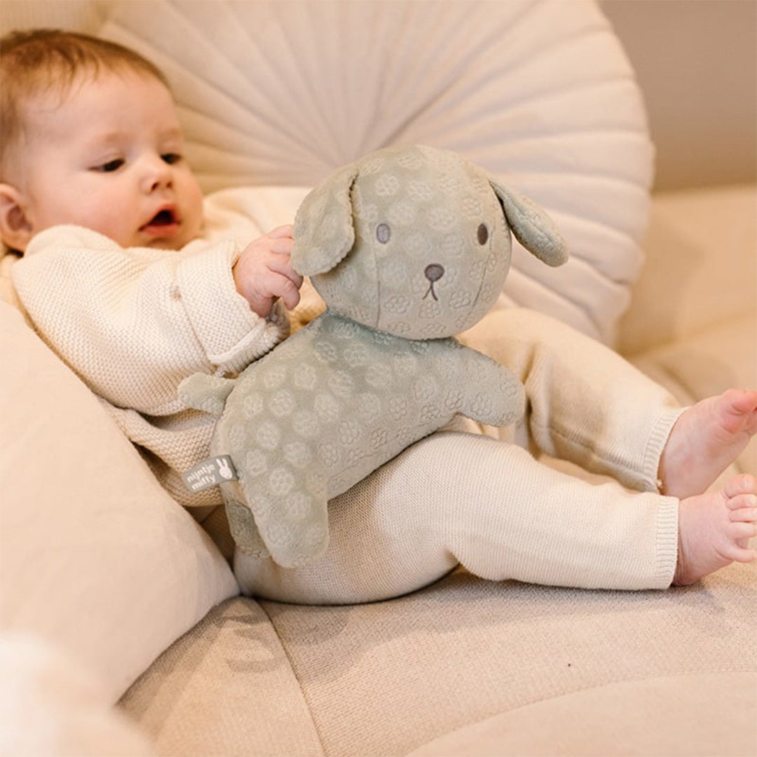 Een baby ligt op een bank en houdt de groene nijntje-miffy pluchen knuffelhond Snuffie vast.