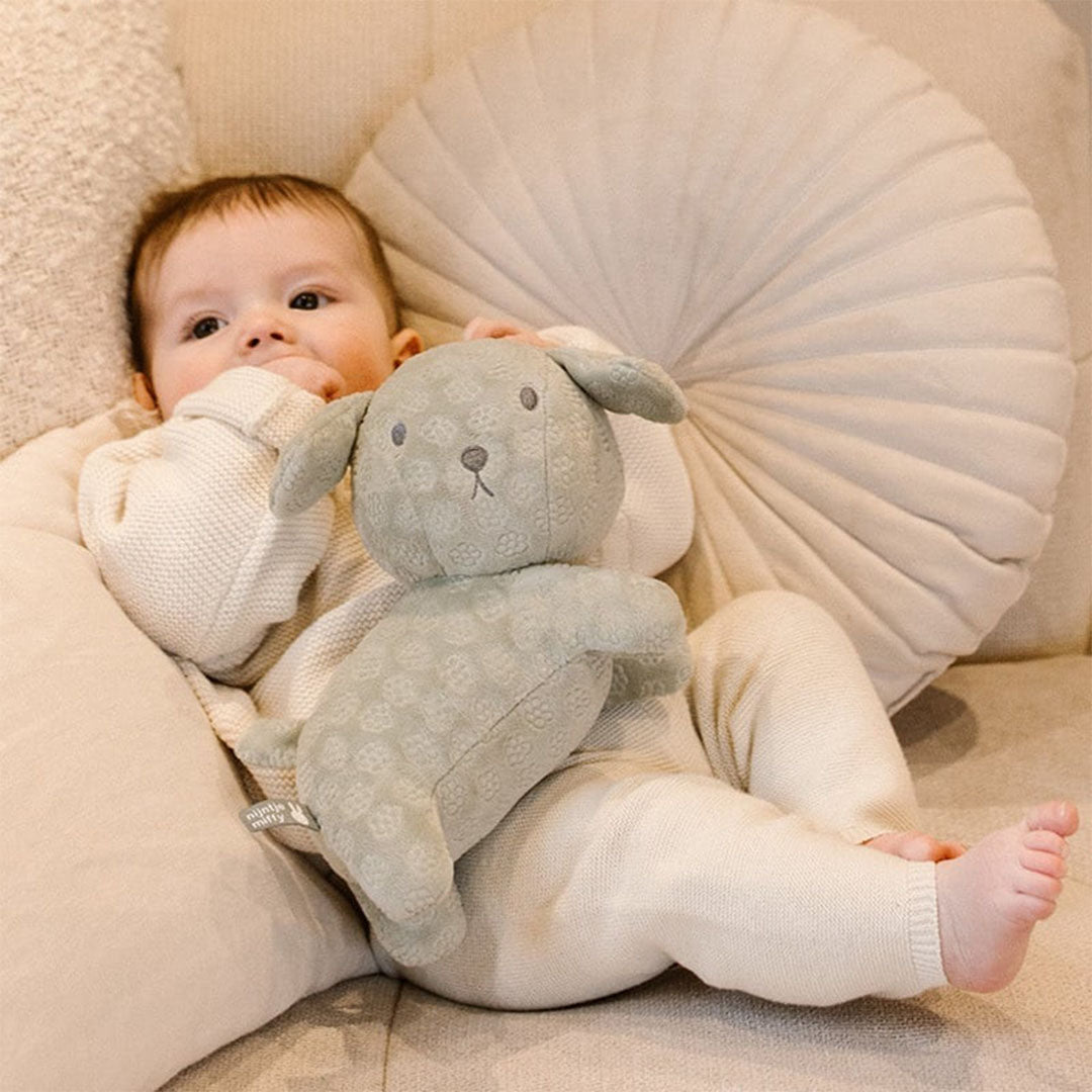 Een baby ligt op een bank en knuffelt met de groene nijntje-miffy pluchen knuffelhond Snuffie.
