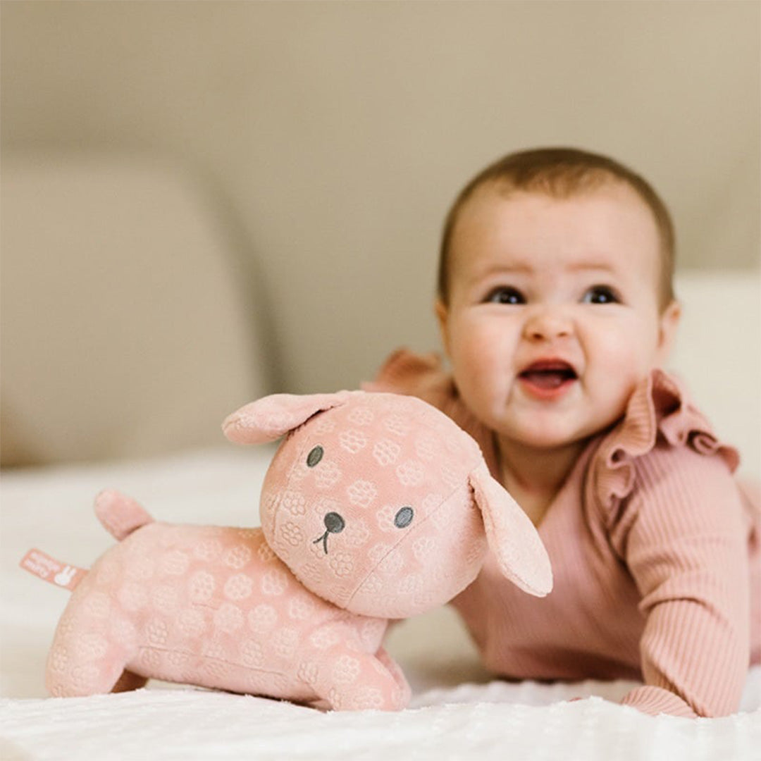 Een lachende baby ligt op een bed naast de Nijntje-Miffy roze pluchen knuffelhond Snuffie.