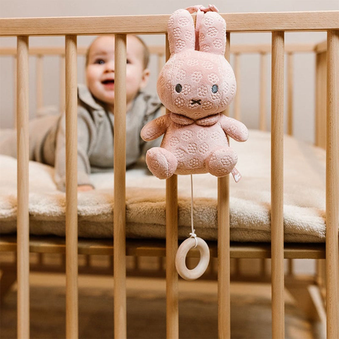 Roze nijntje muziekdoosje met bloemenpatroon hangt aan een houten babybedje, met een lachende baby in de wieg.