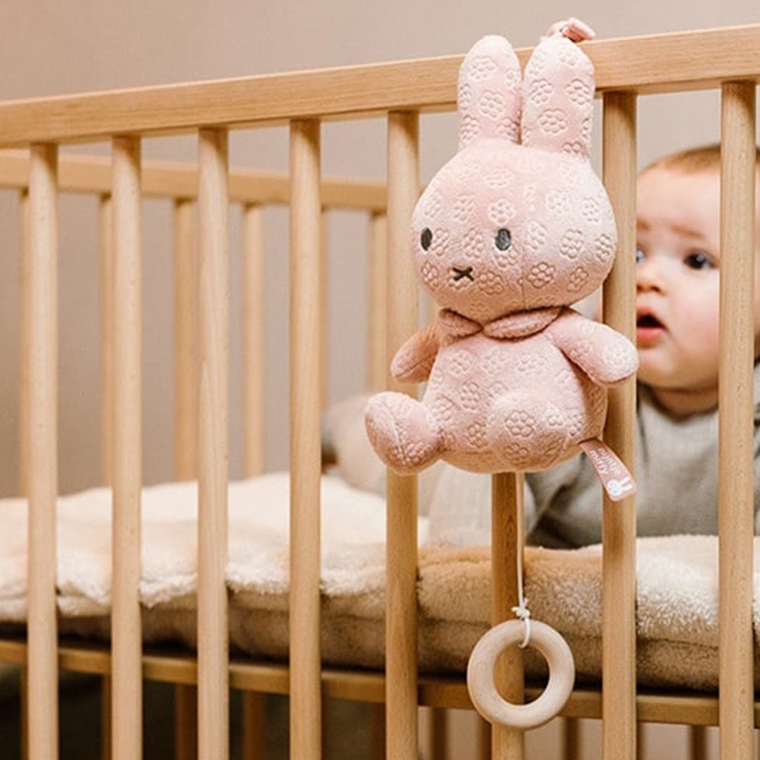 Roze nijntje muziekdoosje met bloemenpatroon hangt aan een houten babybedje, met een baby die ernaar kijkt.