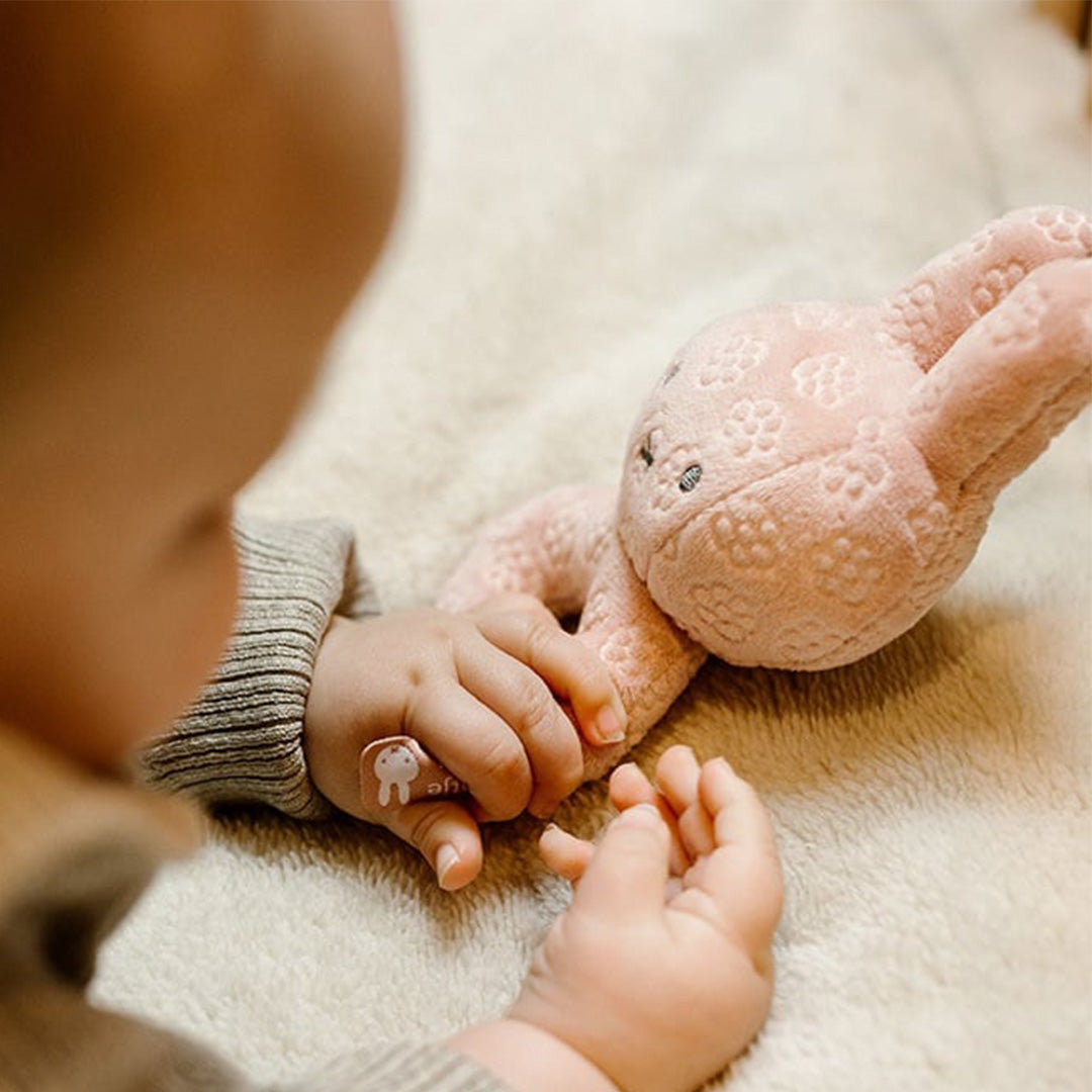 De handen van een baby reiken naar de roze Nijntje rammelaar op een zachte deken.