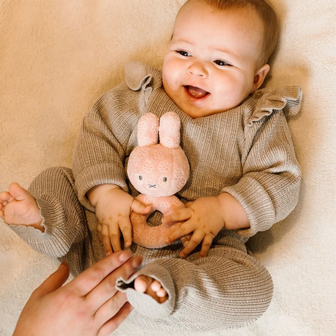 Een lachende baby ligt op de rug en houdt de roze Nijntje rammelaar vast.