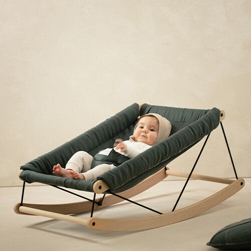 Een baby ligt in een groenblauw nobodinoz babywipstoeltje met houten onderstel.
