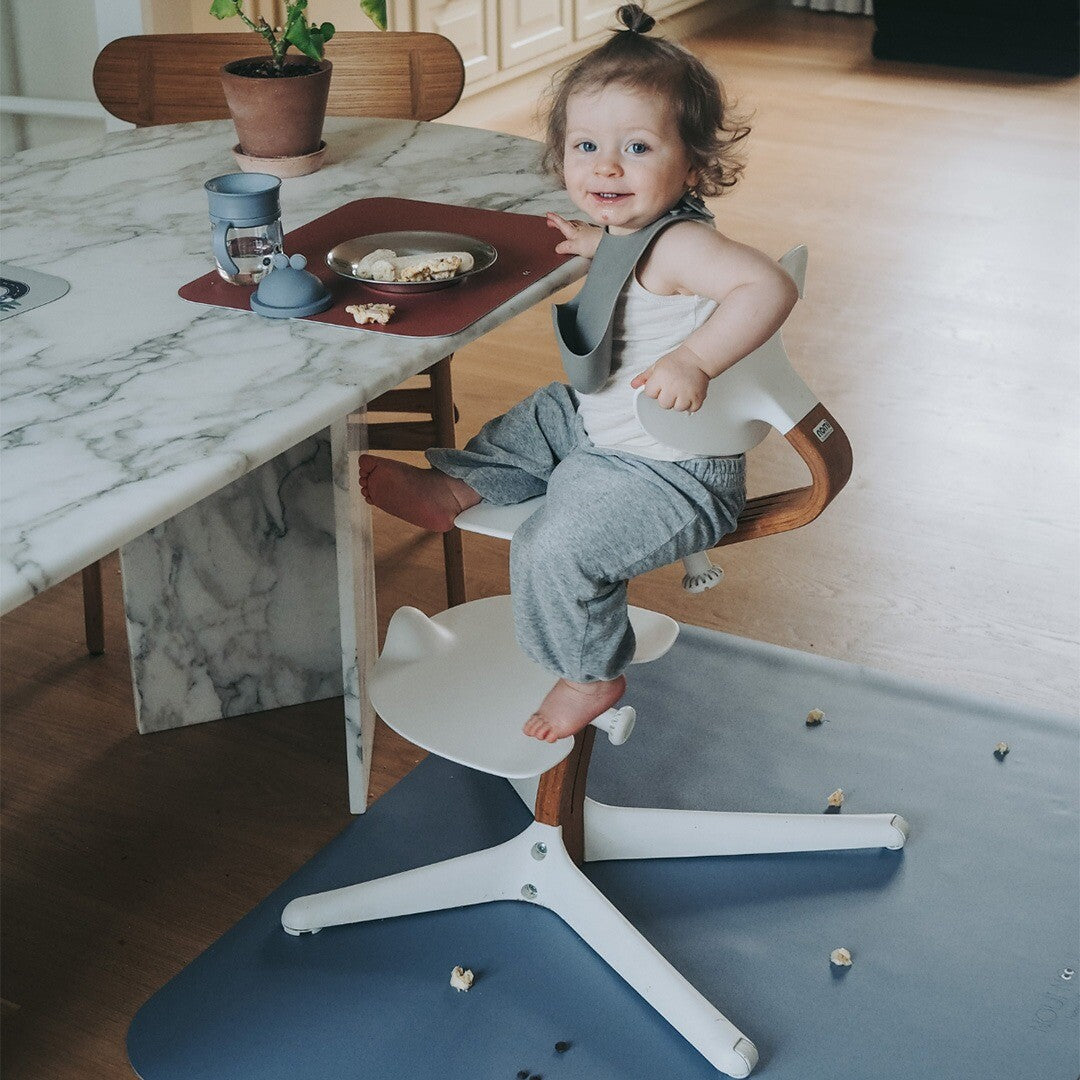 Een baby zit in een kinderstoel met een blauwe noui noui knoeimat op de vloer vol kruimels.