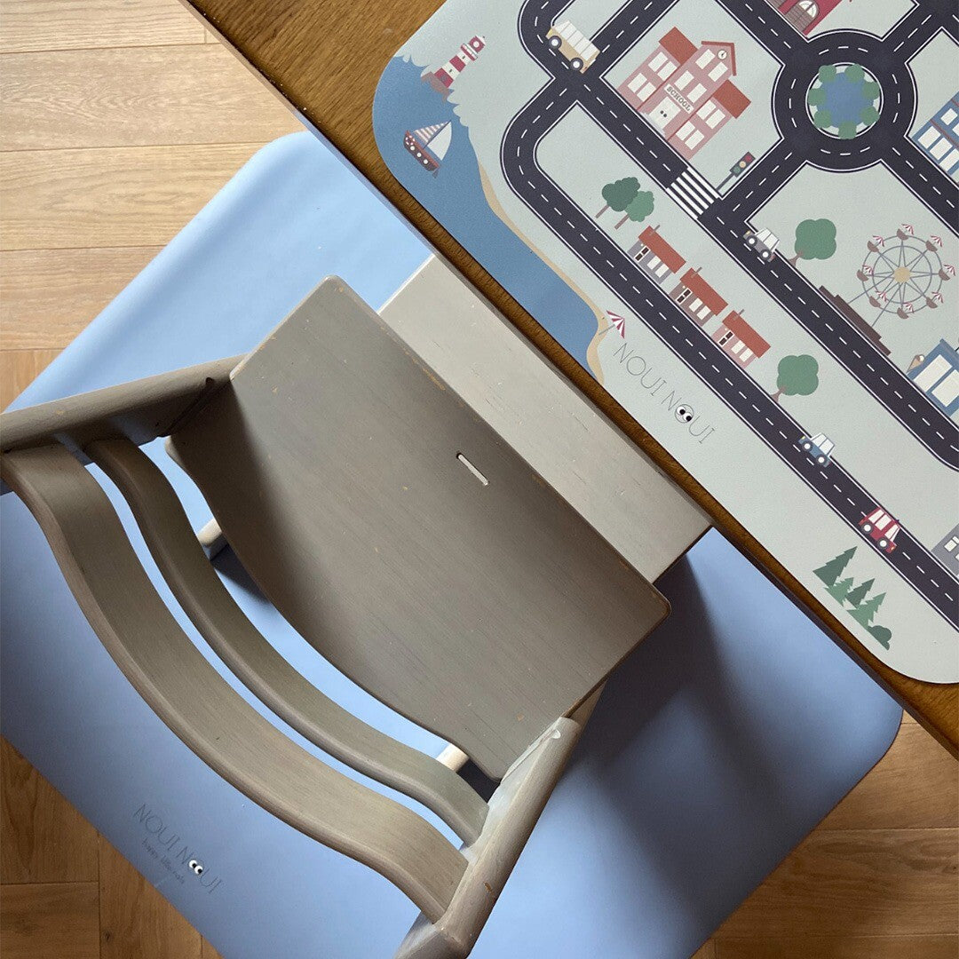 Een lichtblauwe noui noui knoeimat ligt onder een kinderstoel, naast een speelmat met wegenpatroon op tafel.