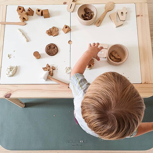 Noui noui knoeimat onder een tafel waar een kind speelt met houten speelgoed en klei.