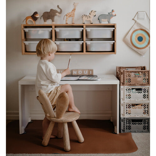 Een kind zit aan een bureau op een houten stoel, met een bruine noui noui knoeimat op de vloer.