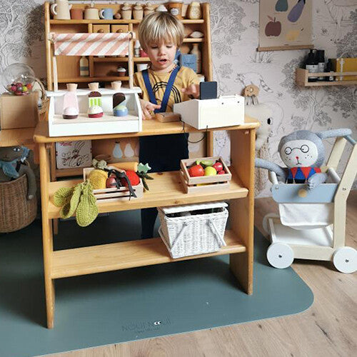 Een kind speelt met een houten speelgoedwinkel, staand op een blauwgroene noui noui knoeimat.