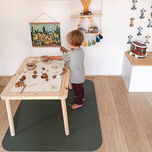 Een kind speelt aan een houten tafel, staand op een donkergroene noui noui knoeimat.
