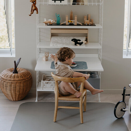 Baby zit op een stoel aan een bureau met een Noui Noui knoeimat op de vloer.