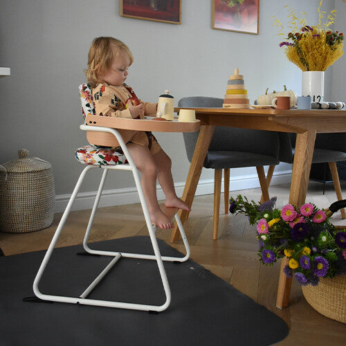 Kind zit in een kinderstoel aan tafel met de Noui Noui knoeimat op de vloer.