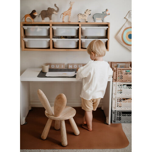 Kind staat aan een bureau met een bruine Noui Noui knoeimat op de vloer.