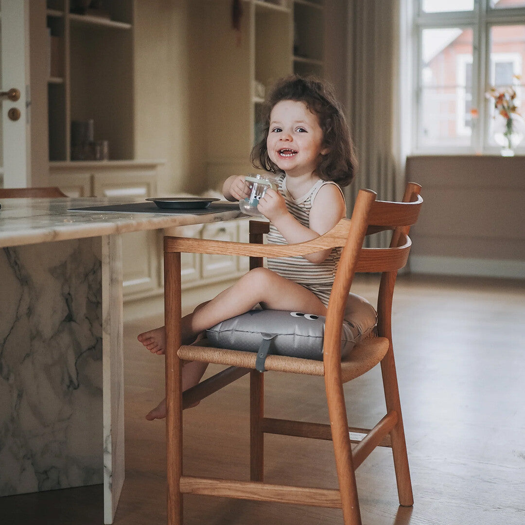 Een lachende peuter zit op het noui noui opblaasbare stoelkussen in een houten kinderstoel aan tafel.