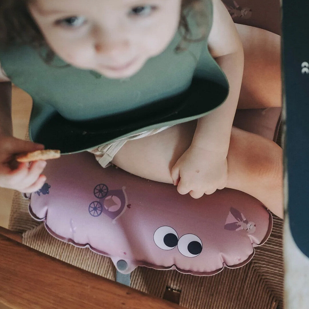 Een peuter zit op het roze noui noui opblaasbare stoelkussen met prinsessenpatroon in een kinderstoel.