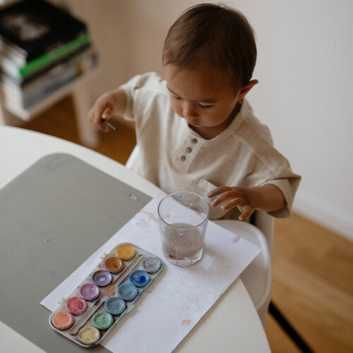 Een baby schildert met waterverf op een lichtgrijze placemat.