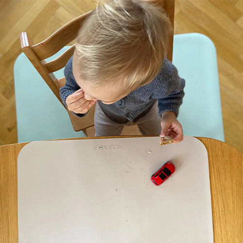 Een peuter eet een snack aan tafel met een lichtbeige Noui Noui placemat en een rood speelgoedautootje.