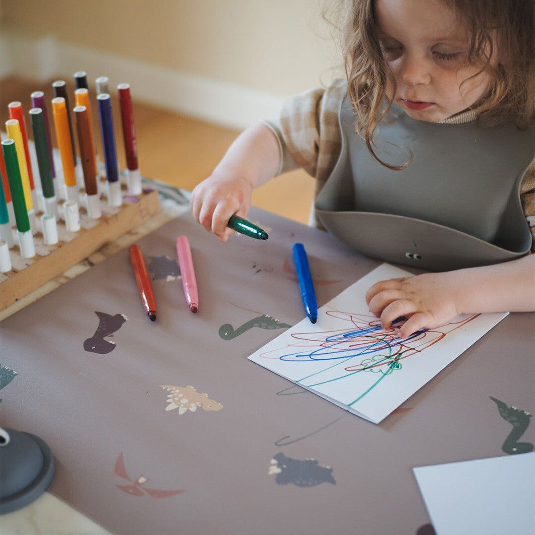 Een kind tekent met kleurpotloden op een wit vel papier dat op de dinosaurus placemat ligt.