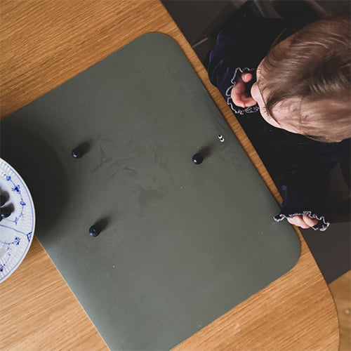 Een babyhandje reikt naar bosbessen op een dusty olive groene placemat op een houten tafel.