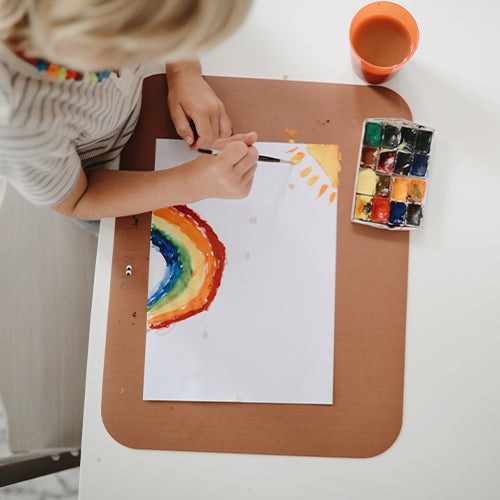 Een kind schildert een regenboog op papier, liggend op een bruine placemat.