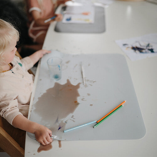 Een kind zit aan tafel met een lichtgrijze noui noui placemat waarop water is gemorst en kleurpotloden liggen.