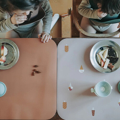 Twee kinderen eten aan tafel met een roze en een bruine noui noui placemat met ijsjespatroon.