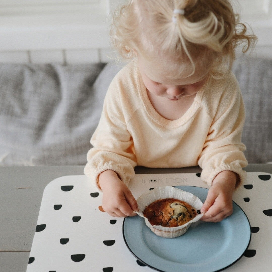 Een kindje pelt een muffin uit op een witte noui noui placemat met zwarte stippen.
