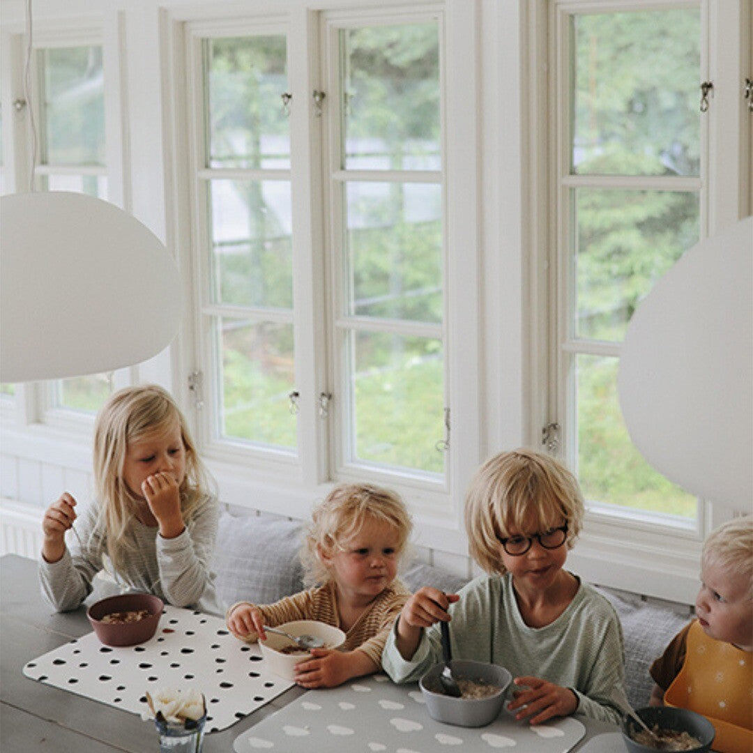 Vier kinderen eten aan tafel, met twee witte noui noui placemats met zwarte stippen en een grijze placemat met wolken.