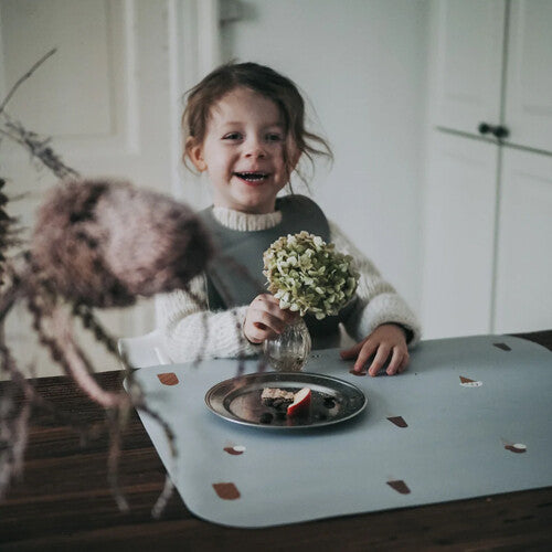 Een lachend kind zit aan tafel met een Noui Noui grijze placemat met ijsjespatroon en een bordje met eten.