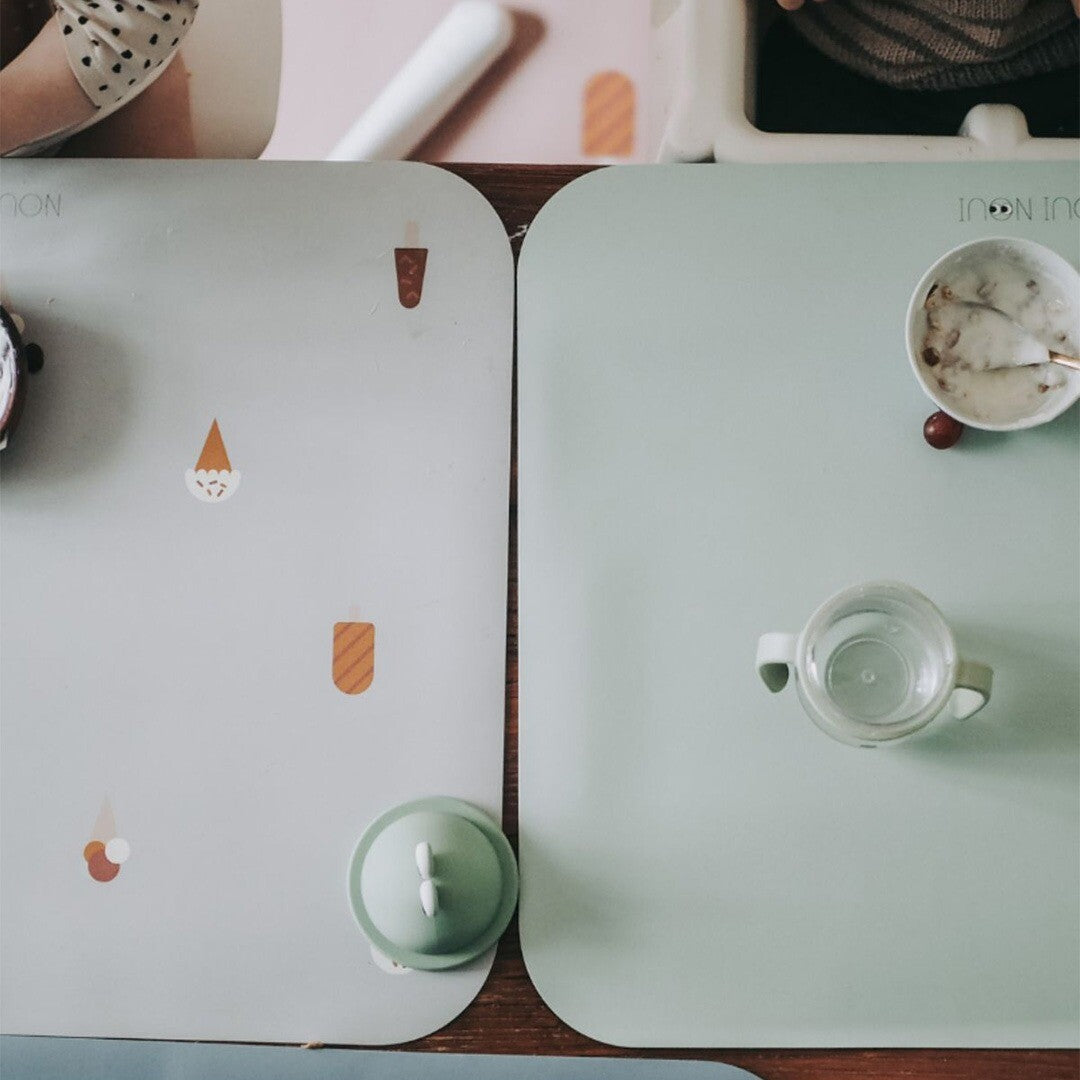 Twee Noui Noui placemats, een grijze met ijsjes en een effen groene, liggen op een houten tafel met kinderkommen en bekers.