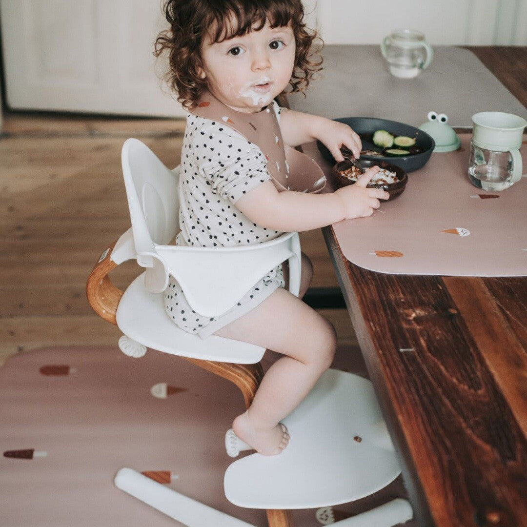 Een kind zit in een kinderstoel aan tafel met een roze Noui Noui placemat met ijsjespatroon.