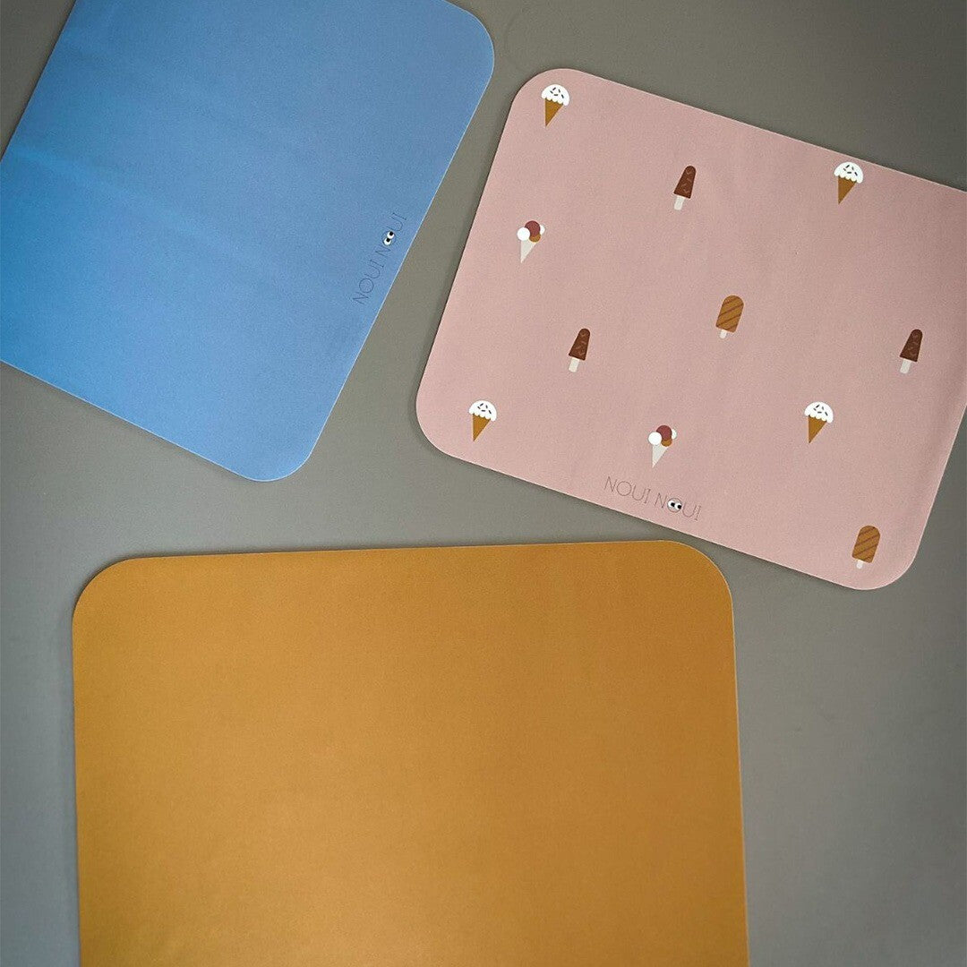 Drie Noui Noui placemats in verschillende kleuren: roze met ijsjespatroon, effen blauw en effen okergeel.