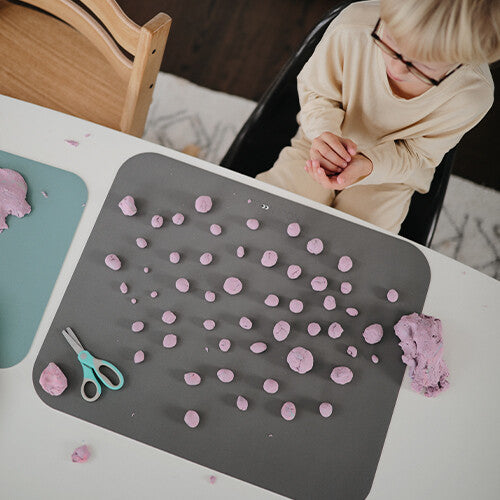 Een donkergrijze placemat met veel kleine paarse kleiballetjes en een schaar.