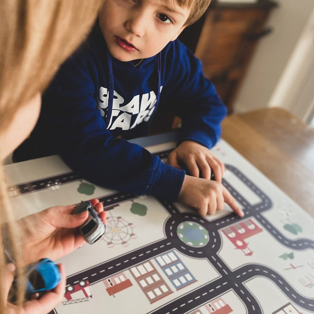Een kind wijst naar de Noui Noui placemat Little Village terwijl een volwassene speelgoedauto's vasthoudt.