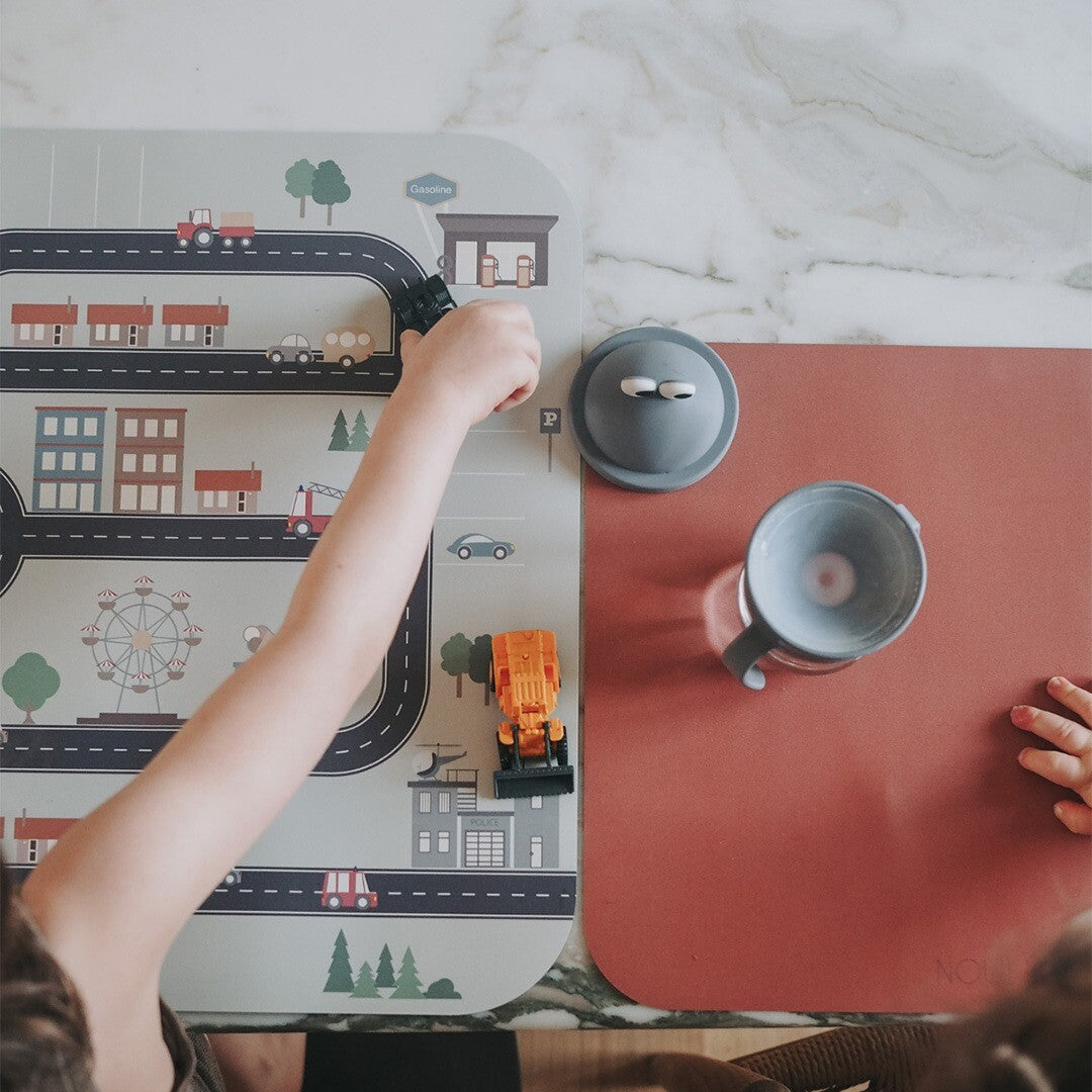 Een kind speelt met een graafmachine op de Noui Noui placemat Little Village naast een roze placemat met een beker.