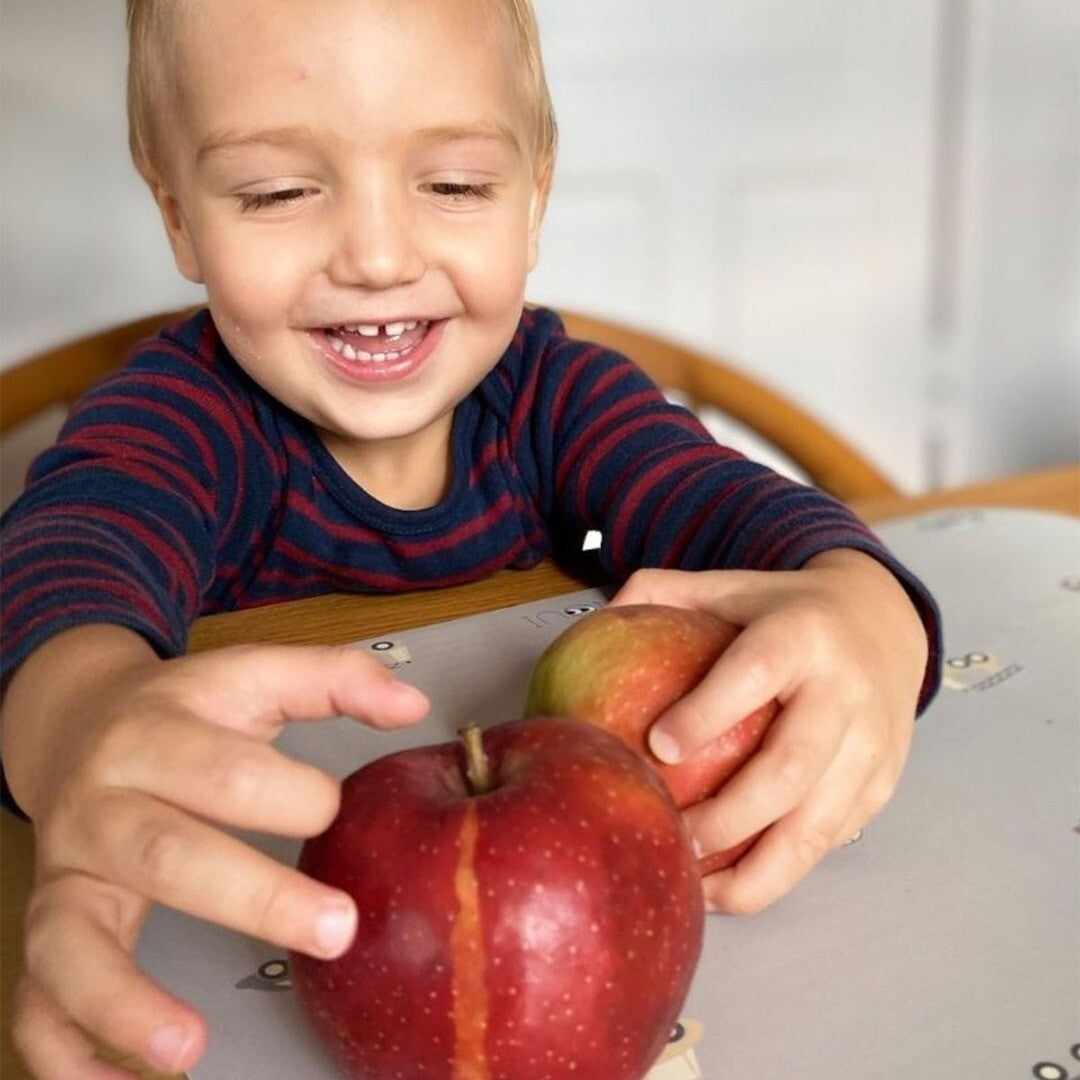 Een lachende peuter met twee appels op een blauwe placemat met voertuigpatroon.
