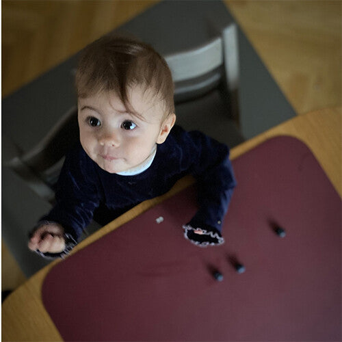 Baby in kinderstoel kijkt omhoog vanaf een donkerrode placemat op een houten tafel.