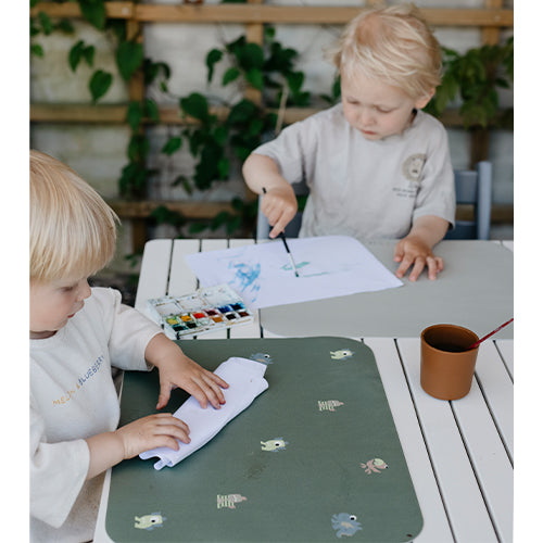 Twee kinderen schilderen en knutselen aan tafel met groene en lichtgrijze placemats.