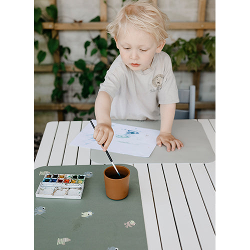 Een kind schildert op papier dat op een lichtgrijze placemat ligt, met een groene placemat ernaast.