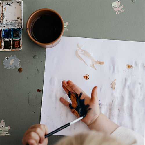 Een kinderhand met verf en een kwast op een groen noui noui placemat met schilderspullen.