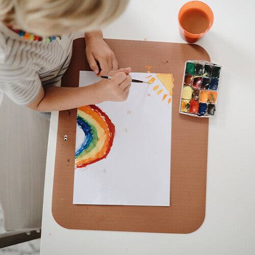 Een kind schildert een regenboog op papier dat op een bruine placemat ligt.