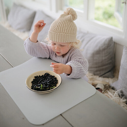 Een peuter met een gebreide muts zit aan tafel met een kom bosbessen op een lichtgrijze placemat.