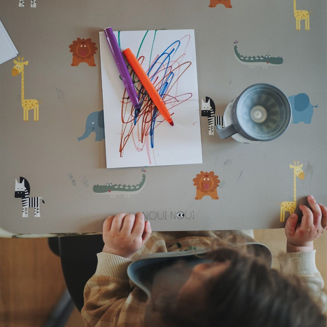 Kind aan tafel met de Noui Noui placemat met safari dierenpatroon, waarop een tekening en stiften liggen.