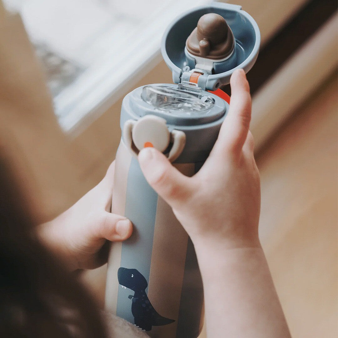 Kinderhandjes openen de noui noui thermosfles met dino-opdruk en gestreepte patroon.