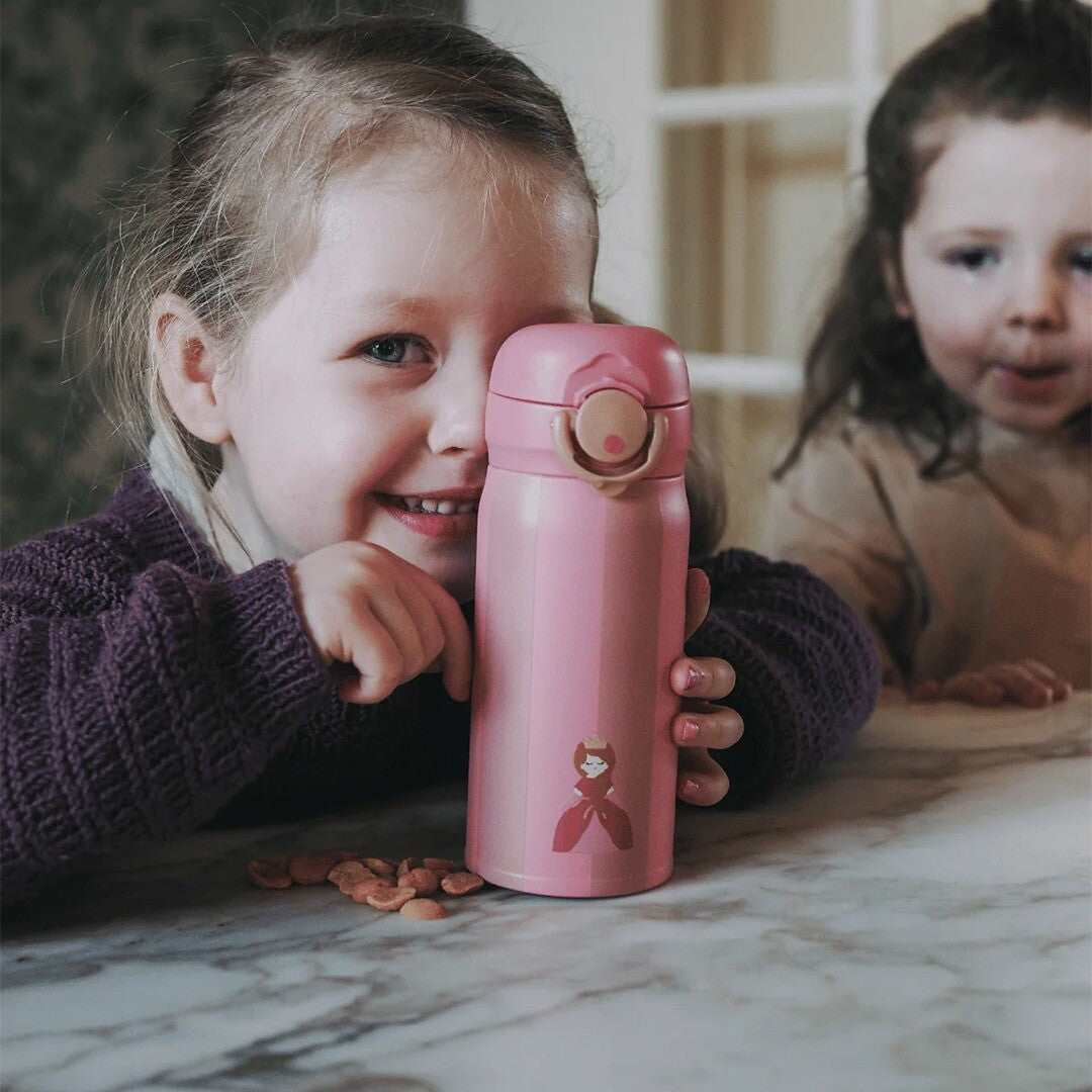 Een lachend kind houdt de roze gestreepte noui noui rvs thermosfles met prinsesfiguur vast aan tafel.