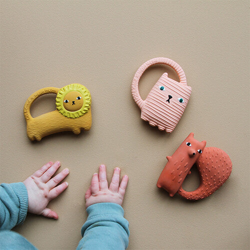 Drie Oli & Carol dieren bijtspeeltjes, een roze kat, oranje eekhoorn en gele leeuw, liggen naast babyhandjes.